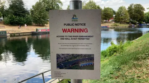 Emma Howgego/BBC A sign saying "public notice warning" in big letters. Further down in smaller writing it reads "Access to the river embankment and wall is not permitted". In the background is a river with grassy banks. Narrowboats are moored on the opposite side of the river