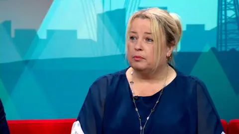 Amanda Hopgood sits on the red Politics North sofa with the teal set behind her. She's wearing a silky navy blue top and has her blonde hair tied back. 