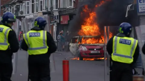 The backs of three police officers are visible as they stand a safe distance from a burning red car on a street. A row of shops can be seen in the distance. A group of people stand beyond the blazing car.