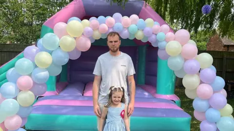 James Turner with short dark hair and beard wearing a grey t-shirt stands behind his daughter Emilee who has dark shoulder length hair and is wearing a blue dress and red rosette. They are standing in front of a multi-colour bouncy castle with an arch of balloons