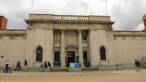 A prestige sandy coloured building with two pillars at the front and two rectangular drapes hanging down from each side of it. You can see people walking along the path and a few people sat in front of the building.
