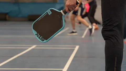Jamie Niblock/BBC A hand holding a pickleball paddle, face open. The paddle has a light blue rim, and a black surface where the polymer ball strikes. There are players, mid-match, in the background. 