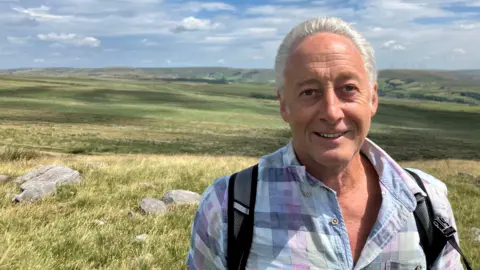 Photograph of Stuart Davies from the Rooley Moor Neighbourhood Forum. The picture is taken on the Cotton Famine Road above Rochdale. He has swept-back grey hair and is wearing an open-necked checked shirt in shades of blue, green and purple.