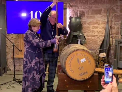 A man and a woman draw whisky out of a barrel and put it into a bottle