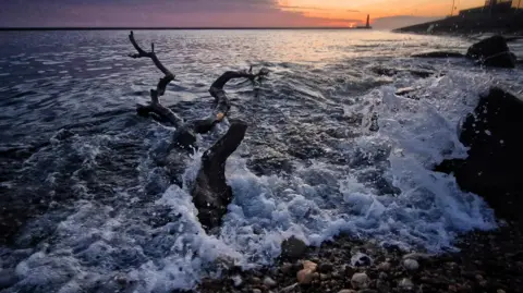 MackemIan The waves are frothy and are thrown into the air as they meet the rocky shore. There is a trunk of wood in the water which is also disturbing the relatively calm water further back. The sun is rising in the distance, casting a lighthouse in silhouette. 