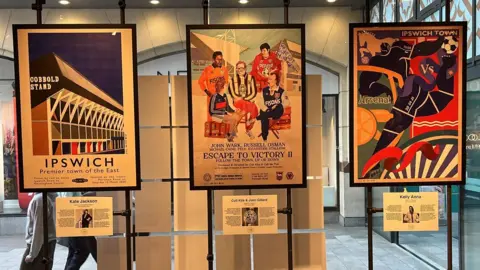 Three of the posters framed and on display in front of a long glass window. All of them have special art designs, all featuring something to do with Ipswich Town Football Club, including players, crests and the Portman Road stadium.