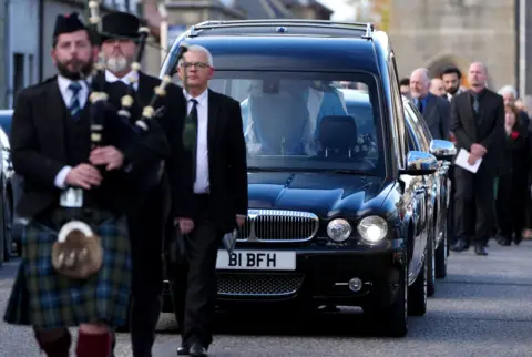 PA Media Salmond's funeral service with the procession coming towards the camera.
There is a male piper in full Highland dress followed by two men in dark suits, white shirts and black ties. The saltire-covered coffin can be seen in the hearse with a mourners' car behind and more people walking in the far distance.