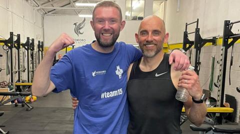 Image of Mark Butcher and David Champion at Kokoro Fitness in Bristol. Mark has dark brown hair and facial hair, and is wearing a blue top. He is flexing his muscles and smiling. David Champion has his arm around Mark. He is wearing a black vest and flexing his muscles too, whilst smilign at the camera. 