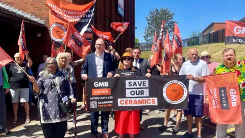 A group of people holding banners and flags. One of the flags reads 'Save Our Ceramics'. The flags bear the GMB union's logo on an orange background.