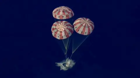 Three red and white parachutes bring the Orion capsule gently into the Pacific Ocean with a splash.