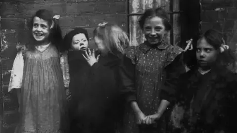 A black and white archive photo of a group of seven young children, most of them girls, on a cobbled street in Belfast in 1926.  Two of the girls are barefoot, one is holding a toddler on her hip.  Two others are hiding in an alleyway. The children are mostly smiling.  A brick building, possibly a large tenement house, is behind them. 