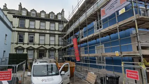 Google The Waverley Hotel in Whitehaven in Cumbria. It is a Georgian-era hotel, with a cream exterior and green-brown window frames. It sits back from the main round, and beside it is a new building under construction. 