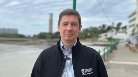 Steve Fitzgibbon standing on the promenade at Havre Des Pas. He has short dark hair. He is wearing a stripey button-up shirt and a "Government of Jersey" zip-up fleece on top.