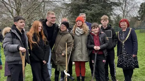 Alice Park Trust A group of young people and adults standing in a line in front of the sapling, some with spades in their hands. 