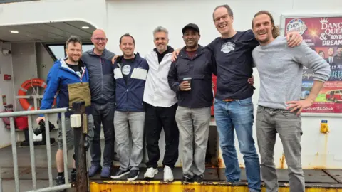 Edmund Phillips A group of seven men stand in a row on a ferry. They have their arms wrapped around one another. They are all smiling and look at the camera.