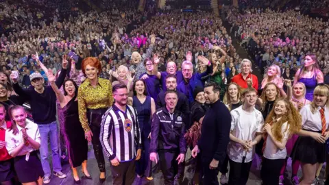John Millard Photography A group of people on stage which the arena audience behind them. Most have their hands up in the air. The people include Matty Healy, Denise Welch, Tim Healy, Stephen Tompkinson and Steph McGovern.