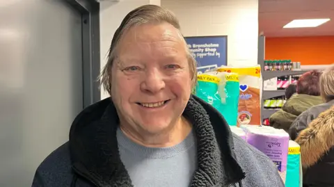 A man in his 70s, with blonde hair combed to one side, smiles as he stands in the shop in front of a shelf full of products. He is wearing a blue and black fleece. A   queue of people can be seen in the background.