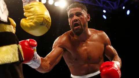 Boxer Lennox Clarke during a match. He is wearing red boxing gloves and has his mouth open as he fights.