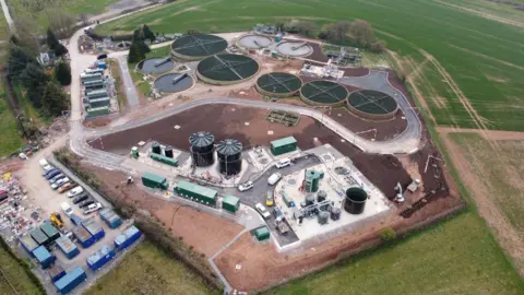 A birds-eye view of the refurbished water recycling centre in Langport. There are round water basins and storage facilities. There is a car park at the edge of the site and around it are green fields.