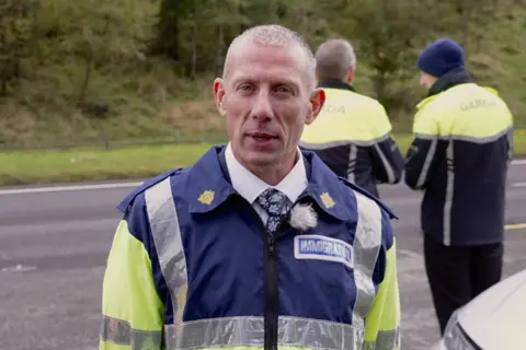 Det Ch Supt Aidan Minnock, with abbreviated  grey hairsbreadth  and wearing a bluish  and yellowish  fluorescent Garda azygous   with an "immigration" label, faces the camera portion    2  different   Garda officers basal   down  him connected  the roadworthy  