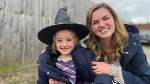 A little girl smiling in a pointy witch hat and blue coat. She is being held by her smiling mother, who has shoulder length blonde hair and is wearing a blue coat.