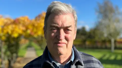 Shaun Whitmore/BBC A picture of Les Garrett. He is standing in his vineyard and smiling at the camera. He has short grey hair and is wearing a blue and grey chequered coat on top of a blue shirt. 