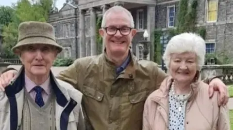 Keith has his arms around his parents as they smile for a photo in front of Mount Stewart. He is wearing a greeny brown bomber jacket and his father on his right is wearing a pink shirt, blue tie and cream coat and bowler hat. His mum on his left is wearing a pink coat and white black dotted top. She has short white hair. 