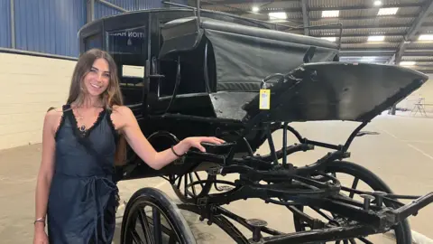 A woman in a dark coloured dress is leaning against an old-fashioned looking carriage. The carriage is black and stands on the floor of a large warehouse with a blue wall to the left hand side.