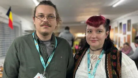 Jon Wright/BBC Albert Arcuda and Cristina Rosu, wearing RCISA lanyards. Albert has a ponytail, glasses and facial hair. He is wearing a khaki green shirt. Cristina is in traditional Romanian dress. She has dyed red and black hair.