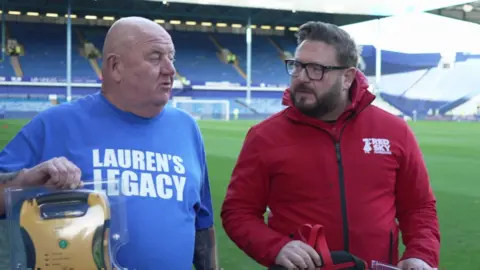 A man wearing a blue t-shirt with the words 'Lauren's Legacy' holds up a defibrillator in its plastic box. Next to him is a bespectacled man wearing a red zipped hoodie.