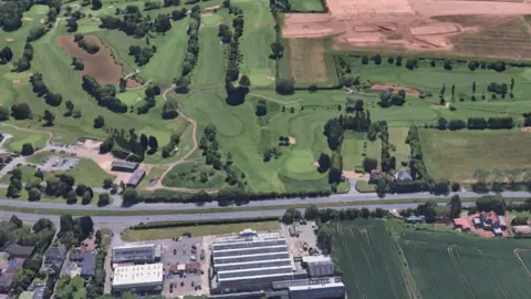 Google An aerial view that shows mostly green fields lined with trees. To the bottom of the image some housing can be seen, as well as some larger buildings and a car park