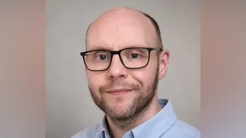 Supplied A male wearing a light blue shirt with the top button undone and black rimmed glasses, looking directly at the camera and smiling slightly. The lower part of his face is covered by stubble, and he is mostly bald on the top of his head. He has a friendly appearance. 