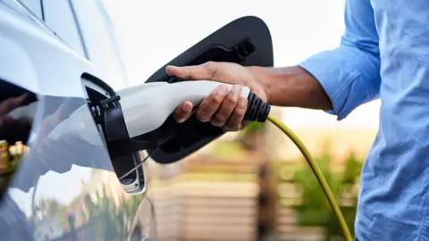 Getty Images Midsection of man plugging in cable while charging electric car.
