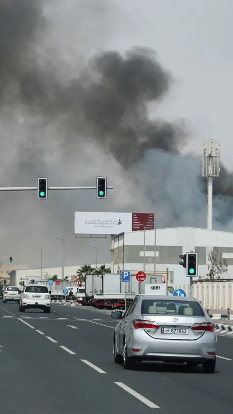 Cars drive down a road in Qatar towards a large billowing cloud of grey smoke coming from a missile strike