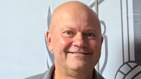 A bald man with blue eyes, wearing a grey top, smiles at the camera.