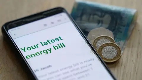 A letter which is being read on a mobile phone. The phone is laid on a wooden table with two one pound coins, a 50p and a £5 note. On the phone it says "Your latest energy bill".