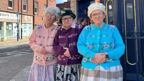 Three women are standing together on a high street wearing colourful knitted cardigans with their hands folded on top of their stomach. They are all wearing glasses and have grey wigs on their heads.