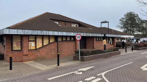 John Devine/BBC Single story building, opened in 1982 as a pub, it has dark roof tiles and reddish brickwork, there is a metal pub signpost outside, with no sign and road markings on a reddish block paved roadway in the foreground