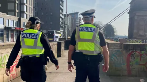 A male and female police officer walking down Clyde Street in Glasgow city centre with their backs to the camera. There is graffiti on the walls and a suspension bridge in the background.