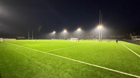 Cinderford's artificial rugby pitch, as seen from one corner. Two sets of posts are visible. It is night time and the pitch is lit by four double floodlights on the near and far sides.