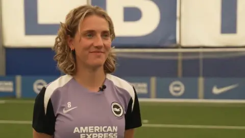 Albion captain Maisie Symonds wears a blue, purple and white club training shirt. A football pitch and advertising hoarding are in the background.