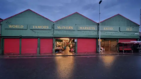 Mangela Coia A row of three large sheds. Along the top of the sheds about red shuttered doors are signs reading: "World famous McIver's markets. Estd. 1921". Two of the doors are open and light spills on to a wet road.