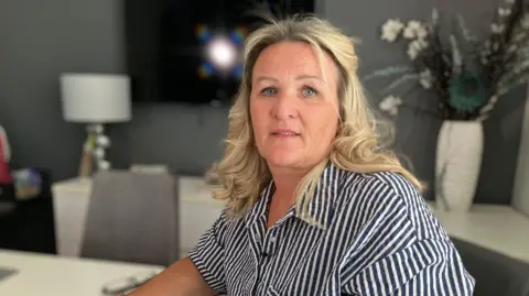 Joanne Hull, wearing a black and white striped blouse. There are flowers in a vase and a table and chairs in the background.