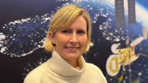 Janine Machin/BBC This is a head and shoulders picture of Kata Escott. She is in the centre of the frame, smiling and looking at the camera. Behind her is a satellite image of the earth from space at night, with lights visible on the land masses. An image of a satellite can also be seen on the wall behind her. 