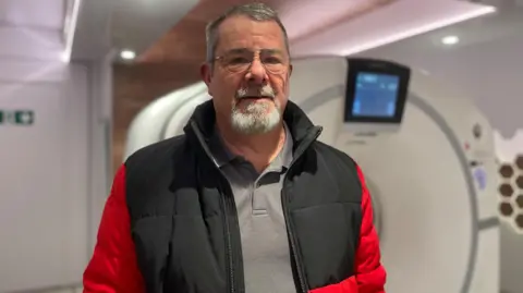 Simon Thake An elderly man with grey hair and beard and a red jacket stands infront of a scanning machine