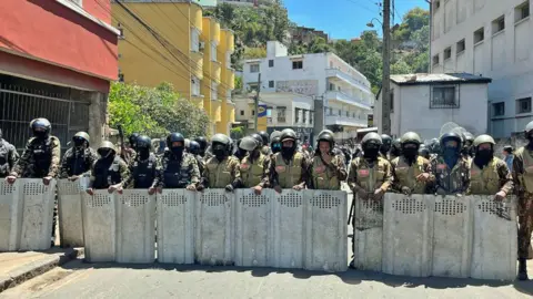 Sammy Awami/BBC A line of police in Antananarivo stand across the street behind metal shields with colorful houses behind them.