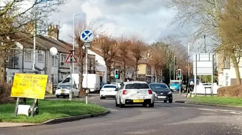 Ellie Calpin A road with cars driving in both directions. There are houses either side. There is a yellow sign reading THIS ROAD WILL BE CLOSED 9 MAR - 15 MAR. It is cloudy above.