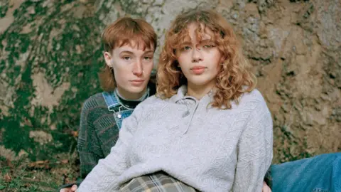 Niamh Shergold Image of the winning portrait called Marina and Connie. It shows two people laying closely together. The pair have light hair and are both wearing knitwear. They are in front of what appears to be a rockface.