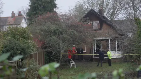 Shaun Whitmore/BBC A back garden and house, with fire officers and a police officer standing close to the building, which has yellow police tape around it. The roof and bricks on the first floor are blackened