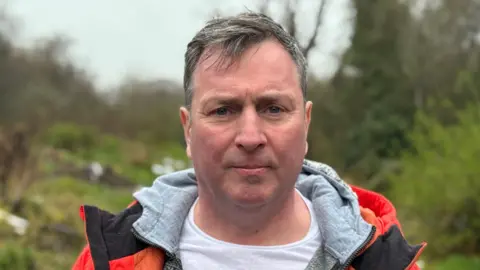 A man in a reflective jacket frowning at the camera while standing in a countryside setting